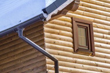 Close-up of corner of new wooden warm ecological cottage house roof covered with snow with steel gutter rain system. Professional construction and drainage pipes installation and connection concept. clipart