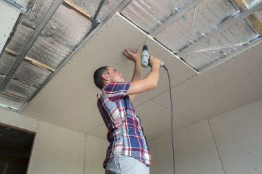 Her zamanki giyim ve iş eldivenleri Alçıpan sabitleme genç adam metal çerçeve parlak alüminyum folyo ile yalıtımlı tavanda Elektrikli tornavida kullanarak tavana askıya. DIY, kendiniz yapın kavramı.
