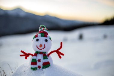 Örgü şapkalı ve eşarplı küçük oyuncak bebek kardan adam. Açık havada, karla kaplı arka planda. Mutlu yıllar ve Mutlu Noeller tebrik kartı teması.