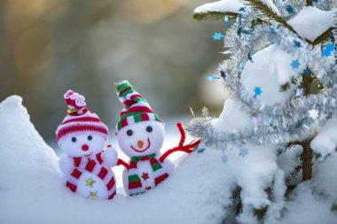 İki komik oyuncak bebek kardan adam örgü şapkalı ve eşarplı açık havada, çam ağacı dalının yanında. Mutlu yıllar ve Mutlu Noeller tebrik kartı.