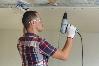 Alçıpan sabitleme Gözlük Genç adam metal çerçeve parlak alüminyum folyo ile yalıtımlı tavanda Elektrikli tornavida kullanarak tavana askıya. Tadilat, inşaat, do It yourself konsepti.