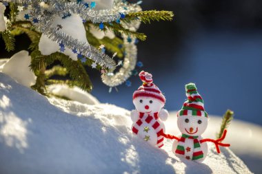 İki komik oyuncak bebek kardan adam örgü şapkalı ve eşarplı açık havada, çam ağacı dalının yanında. Mutlu yıllar ve Mutlu Noeller tebrik kartı.
