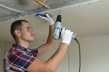 Alçıpan sabitleme Gözlük Genç adam metal çerçeve parlak alüminyum folyo ile yalıtımlı tavanda Elektrikli tornavida kullanarak tavana askıya. Tadilat, inşaat, do It yourself konsepti.