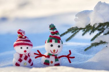 İki komik oyuncak bebek kardan adam örgü şapkalı ve eşarplı açık havada, çam ağacı dalının yanında. Mutlu yıllar ve Mutlu Noeller tebrik kartı.