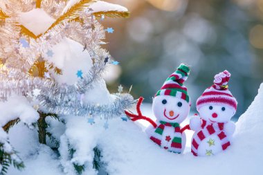 İki komik oyuncak bebek kardan adam örgü şapkalı ve eşarplı açık havada, çam ağacı dalının yanında. Mutlu yıllar ve Mutlu Noeller tebrik kartı.