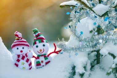 İki komik oyuncak bebek kardan adam örgü şapkalı ve eşarplı açık havada, çam ağacı dalının yanında. Mutlu yıllar ve Mutlu Noeller tebrik kartı.