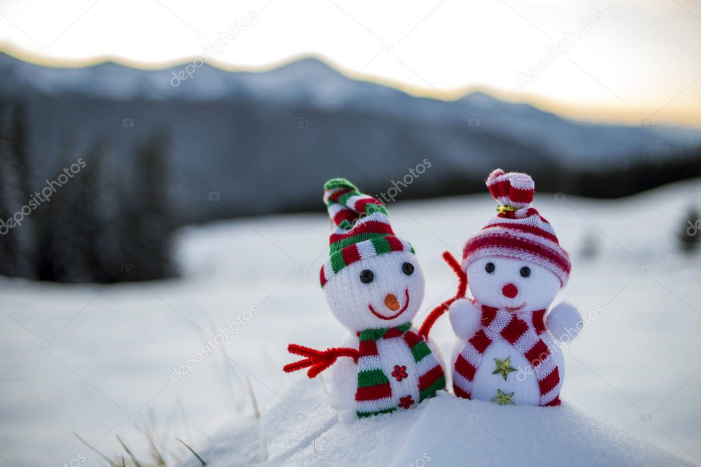 Two small funny toys baby snowman in knitted hats and scarves in deep snow outdoors on blurred mountains landscape background. Happy New Year and Merry Christmas greeting card theme.