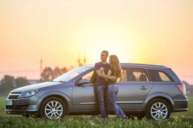 Genç çift aşk, çekici ince uzun - saçlı kadın ve yakışıklı stand yeşil alanda parlak gökyüzü günbatımı veya gündoğumu kopya alan arka plan, gümüş renkli araba, kucakladı birlikte yaslanmış.