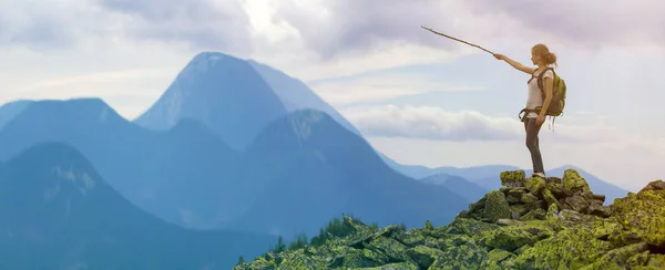 Young slim blond tourist girl with backpack points with stick at foggy mountain range panorama standing on rocky top on bright blue morning sky background. Tourism, traveling and climbing concept.