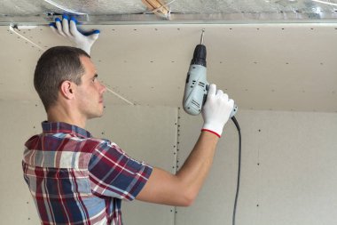 Her zamanki giyim ve iş eldivenleri Alçıpan sabitleme genç adam metal çerçeve parlak alüminyum folyo ile yalıtımlı tavanda Elektrikli tornavida kullanarak tavana askıya. DIY, kendiniz yapın kavramı.