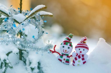 İki komik oyuncak bebek kardan adam örgü şapkalı ve eşarplı açık havada, çam ağacı dalının yanında. Mutlu yıllar ve Mutlu Noeller tebrik kartı.