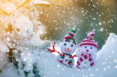 İki komik oyuncak bebek kardan adam örgü şapkalı ve eşarplı açık havada, çam ağacı dalının yanında. Mutlu yıllar ve Mutlu Noeller tebrik kartı.