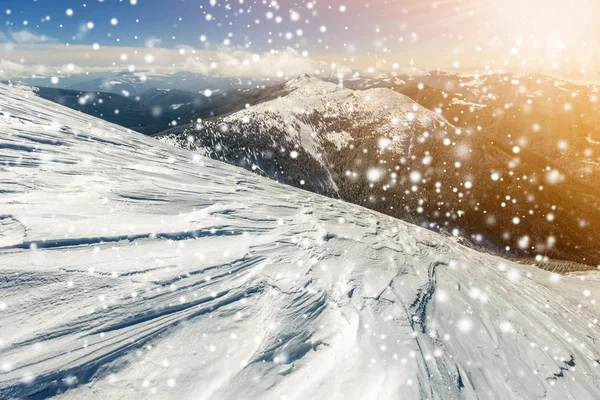 Beautiful winter landscape. Steep mountain hill slope with white deep snow, distant mountain range panorama, large snowflakes and bright shining sun rays on blue sky colorful copy space background.