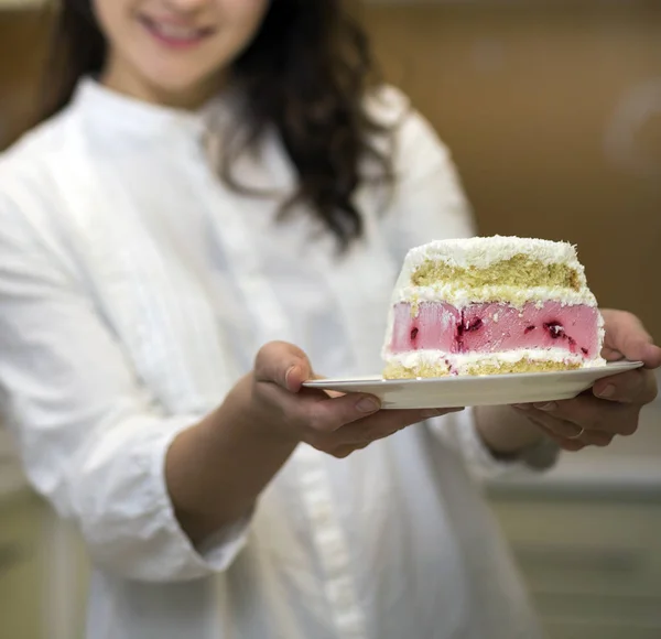 Young attractive smiling woman holding plate with fresh delicious yummy homemade piece of fruit biscuit white and pink creamy cake on blurred background.