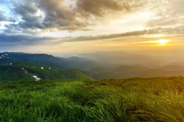 Geniş Yataklı güneş doğarken yaz. Parlayan turuncu güneş mavi bulutlu gökyüzü yeşil çimenli tepe yumuşak çim ve uzak dağ üzerinde yükselterek sabah sis ile kaplı. Doğa kavramının güzellik.