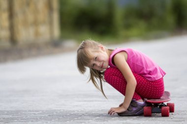 Genç ve güzel uzun saçlı sarışın çocuk kız rahat pembe giyim bulanık yaz yeşil zemin üzerine güneşli sokakta kaykay üzerinde ellerinde yaslanmış oturuyor. Çocuk Aktiviteleri, Oyunlar ve eğlence kavramı.