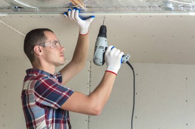 Alçıpan sabitleme Gözlük Genç adam metal çerçeve parlak alüminyum folyo ile yalıtımlı tavanda Elektrikli tornavida kullanarak tavana askıya. Tadilat, inşaat, do It yourself konsepti.