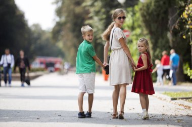 Mutlu aile, geri tutuşup iki sevimli çocuk, kızı ve oğlu ile güneşli park sokak yeşil ağaçlar bokeh arka plan üzerinde üzerinde sıcak yaz gününde arayan genç sarışın uzun saçlı kadın arkadan görünüm.