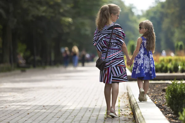 Genç sarışın uzun saçlı ince çekici kadın ve kız küçük çocuk güneş gözlüğü ve şık elbiseler güneşli yaz park uğrayacağız tutuşup yürürken görünümünü geri. Mutlu aile ilişkileri kavramı.