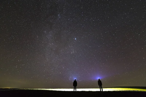 Back view of two men with head flashlights on green grassy field under beautiful dark blue summer starry sky. Night photography, beauty of nature concept. Wide angle shot, copy space background.