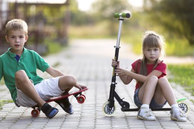 İki sevimli mutlu komik gülümseyen genç çocuk ve kamera için poz scooter ve güneşli renkli arka plan bulanık üzerinde kaykay üzerinde oturan güzel uzun saçlı kız yakışıklı çocuk kız.