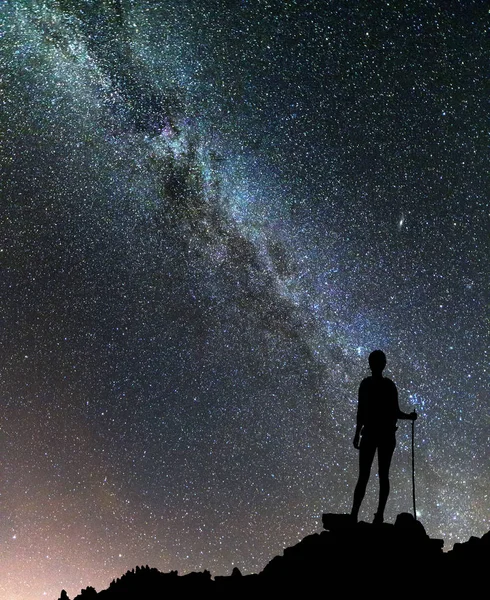 Back view of young slim backpacker tourist girl on rocky mountain top on dark night starry sky and foggy mountain range panorama background. Tourism, traveling and climbing concept.