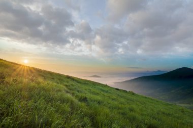 Dağ manzarası gündoğumu, güzel havalarda. Yeşil çimenli sarp bir tepe, sisli Vadisi ve ile parlak mavi gökyüzü altında uzak dağ Güneş beyaz bulutlar yükselterek yaktı. Doğa kavramının güzellik.
