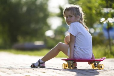 Sevimli küçük sarışın kız beyaz şort ve kaykay üzerinde oturan ve düşünceli bir ışık kamera seyir T-shirt yaz arka plan bulanık. Çocuk Aktiviteleri ve aktif yaşam tarzı kavramı.