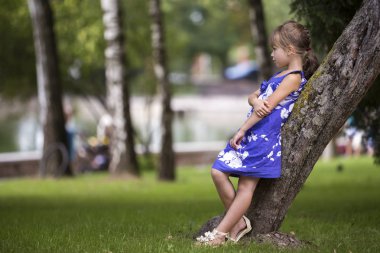 Sevimli Genç ve güzel çocuk kız güzel uzun sarı saçlı, mavi moda elbise sersem park yeşil çimenli bahçede bulanık ağaçlar ve göl arka plan üzerinde gülümseyen büyüyen ağaç gövdesi üzerine eğilerek.