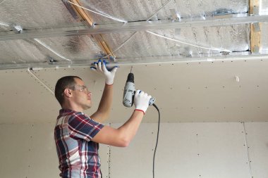 Alçıpan sabitleme Gözlük Genç adam metal çerçeve parlak alüminyum folyo ile yalıtımlı tavanda Elektrikli tornavida kullanarak tavana askıya. Tadilat, inşaat, do It yourself konsepti.