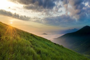 Dağ manzarası gündoğumu, güzel havalarda. Yeşil çimenli sarp bir tepe, sisli Vadisi ve ile parlak mavi gökyüzü altında uzak dağ Güneş beyaz bulutlar yükselterek yaktı. Doğa kavramının güzellik.
