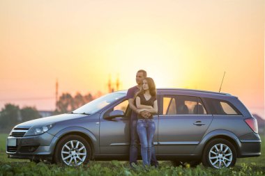 Genç çift, çekici ince uzun - saçlı kadın ve yakışıklı adam kucakladı birlikte gümüş renkli araba yeşil parlak gökyüzü günbatımı veya gündoğumu kopya alan arka plan üzerinde kırsal kırsal alanında, yaslanmış.