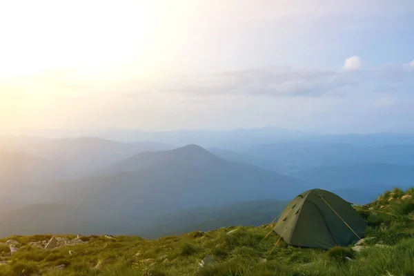 Summer camping in mountains at dawn. Tourist tent on round grassy hill on distant misty blue mountains range under pink sky before sunrise or sunset. Tourism, hiking and beauty of nature concept.