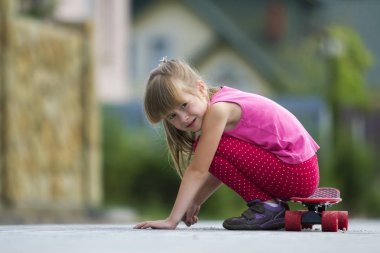 Genç ve güzel uzun saçlı sarışın çocuk kız rahat pembe giyim bulanık yaz yeşil zemin üzerine güneşli sokakta kaykay üzerinde ellerinde yaslanmış oturuyor. Çocuk Aktiviteleri, Oyunlar ve eğlence kavramı.