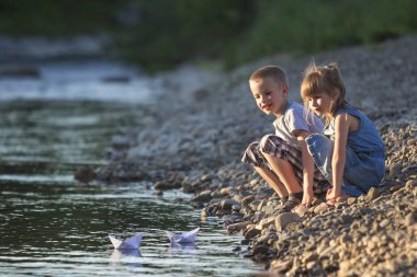 İki sevimli sarışın çocuk, erkek ve kız nehir kıyısında su beyaz kağıt tekne içinde parlak yaz gönderme mavi arka plan bulanık. Sevinçlerini ve mutlu çocukluk ve açık hava faaliyetleri kavramı ile oyun.