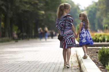 Genç sarışın uzun saçlı ince çekici kadın ve kız küçük çocuk güneş gözlüğü ve şık elbiseler güneşli yaz park uğrayacağız tutuşup yürürken görünümünü geri. Mutlu aile ilişkileri kavramı.