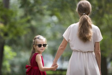 Güneş gözlüğü ve şık kırmızı elbise holding oldukça küçük sarı uzun saçlı çocuk kız arkadan görünüm trustingly el bulanık renkli güneşli yaz bokeh arka plan üzerine açık havada Genç ince kadın.