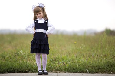 Tam uzunlukta portre sevimli tatlı ciddi düşünceli ilk sınıf öğrencisi kızın okul üniforması ve beyaz yay bulanık ışık yeşil güneşli çimen ve beyaz gökyüzü kopya alan arka plan üzerinde uzun sarı saç.