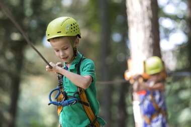 Genç Sevimli sarışın çocuk boy gülümseyerek emniyet kemeri ve karabina ile kablosuna bağlı kask ip yol boyunca parlak güneşli bokeh arka plan üzerinde taşır. Spor, oyun, eğlence, açık hava etkinliği konsepti.