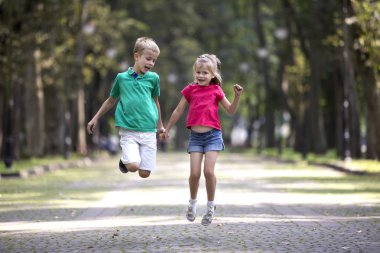Komik çocuk, kız ve erkek gülümseyen iki sevimli genç kız ve erkek kardeşim, atlama ve having fun parlak güneşli park sokak yeşil ağaçlar bokeh arka plan bulanık. Mutlu dikkatsiz çocukluk kavramı.
