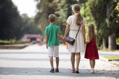 Arkadan mutlu aile, Genç sarışın uzun saçlı kadın iki çocuk, küçük kızı ve oğluyla güneşli park sokak boyunca yürüyen tutuşup görünüşü yeşil ağaçlar bokeh arka plan üzerinde sıcak yaz gününde.