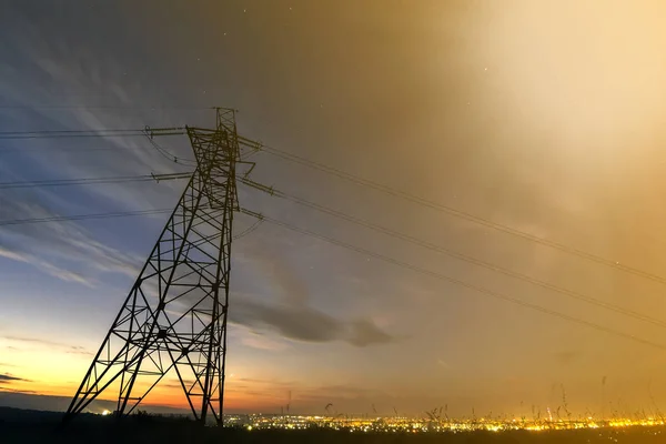 Transmission and long distance distribution of electricity concept. High voltage tower with electric power lines stretching on dark blue starry sky.and bright distant city lights background.