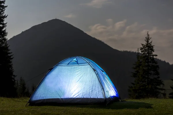 Tourist hikers tent brightly lit from inside on green grassy valley among pine trees under dark blue cloudy sky on distant mountain covered with forest background. Summer night camping in mountains.