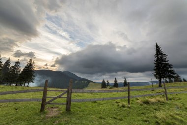 Yeşil çimenli Vadisi yamaç ilkel ahşap çit ve kapı ile panoramik yaz görünümünü gündoğumu, mavi bulutlu gökyüzü altında uzak odunsu sisli dağda. Güzellik, Doğa ve kavram seyahat.