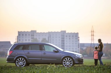 Genç ince çekici uzun saçlı kadın anne ve gümüş renkli araba bulanık apartman ve berrak gökyüzü kopya alan arka plan üzerinde yeşil alanında el ele tutuşarak küçük çocuk kız kızı arkadan görünüm.