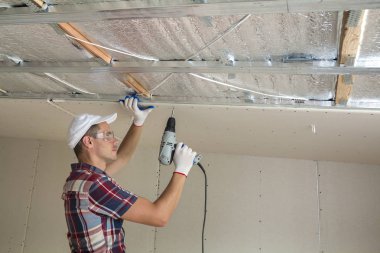 Alçıpan sabitleme Gözlük Genç adam metal çerçeve parlak alüminyum folyo ile yalıtımlı tavanda Elektrikli tornavida kullanarak tavana askıya. Tadilat, inşaat, do It yourself konsepti.