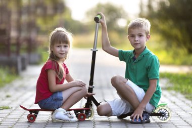 İki sevimli mutlu komik gülümseyen genç çocuk ve kamera için poz scooter ve güneşli renkli arka plan bulanık üzerinde kaykay üzerinde oturan güzel uzun saçlı kız yakışıklı çocuk kız.
