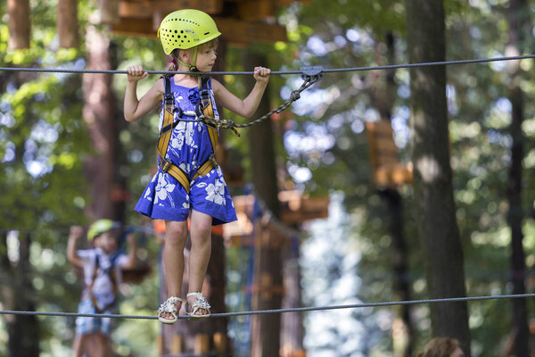 Young child girl in safety harness and helmet attached with carbine to cable moves carefully along rope way in recreation park on green foliage sunny bokeh background. Sport, game and leisure concept.