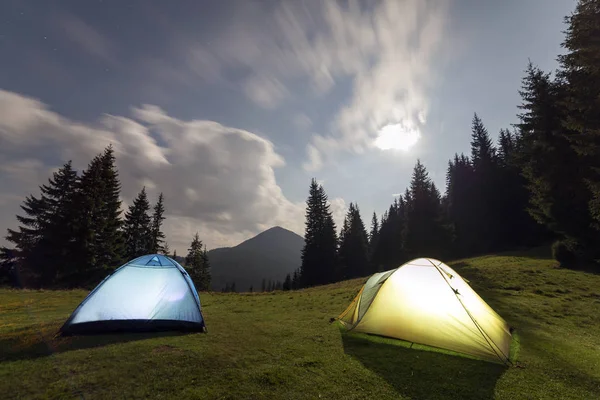 Bright big moon in dark blue cloudy sky over two tourist tents on green grassy forest clearing among tall pine trees on distant mountain background. Tourism, night camping in summer mountains.