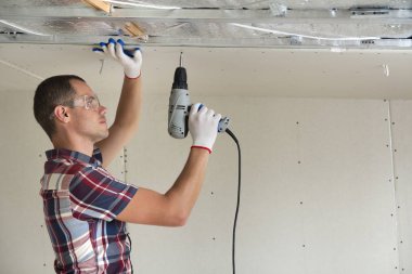 Alçıpan sabitleme Gözlük Genç adam metal çerçeve parlak alüminyum folyo ile yalıtımlı tavanda Elektrikli tornavida kullanarak tavana askıya. Tadilat, inşaat, do It yourself konsepti.